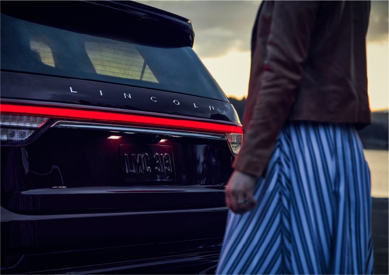 A person is shown near the rear of a 2023 Lincoln Aviator® SUV as the Lincoln Embrace illuminates the rear lights | Bergstrom Lincoln of Neenah in Neenah WI
