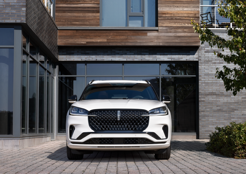 A white 2025 Lincoln Aviator® SUV is shown parked outside a modern building with gray brick and wood accents. | Bergstrom Lincoln of Neenah in Neenah WI