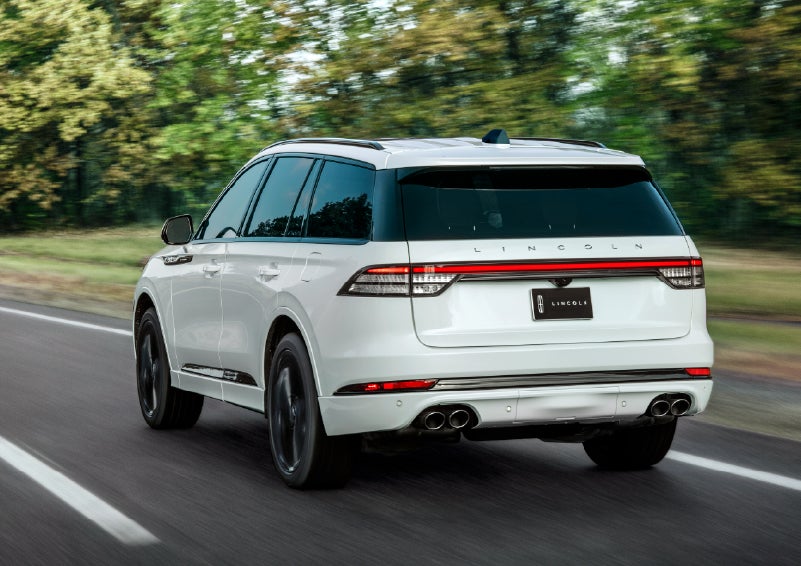 A white 2026 Lincoln Aviator® SUV is being driven away down a road lined with colorful trees. | Bergstrom Lincoln of Neenah in Neenah WI